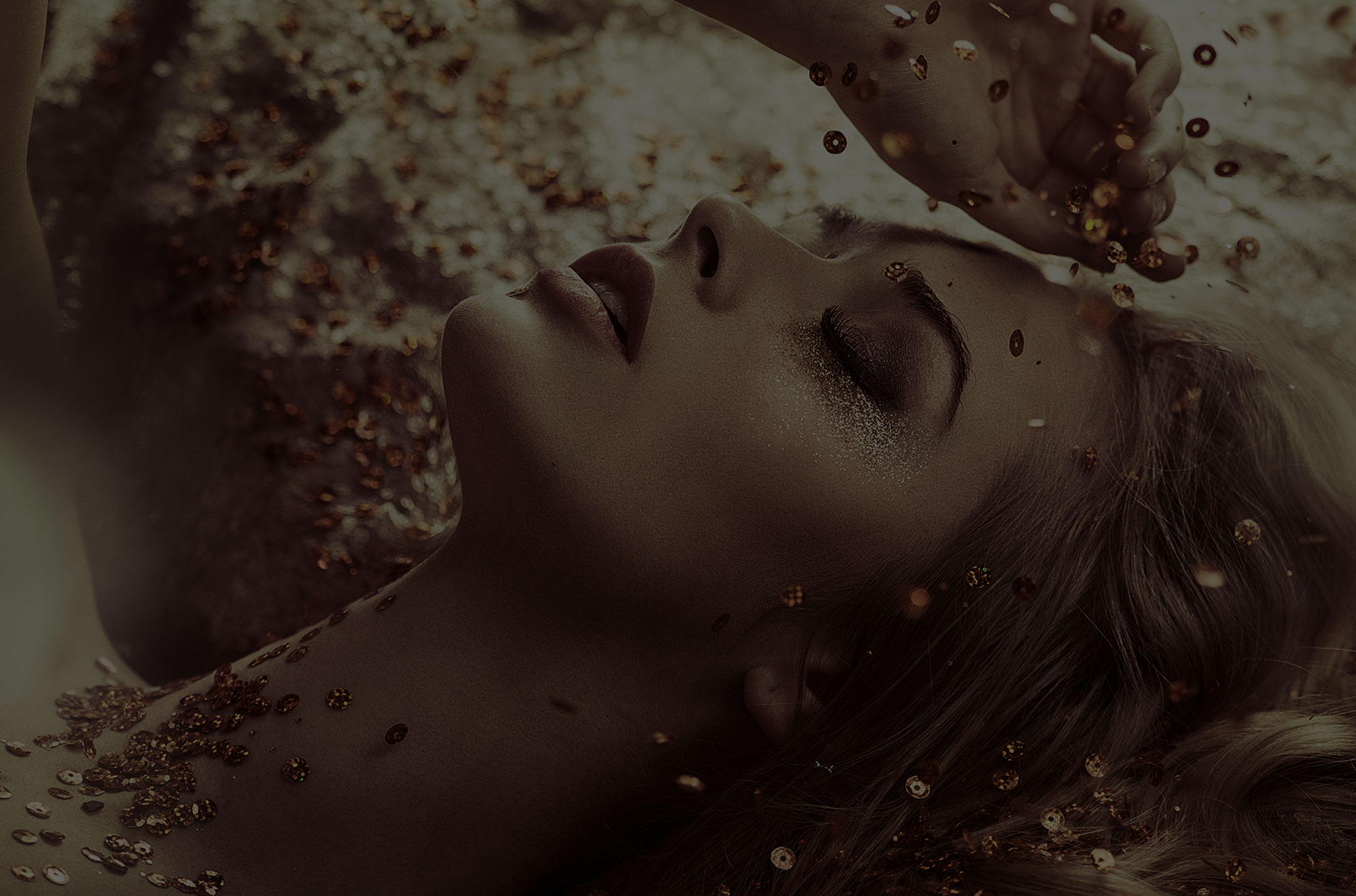 Blonde woman lying back as gold sequins fall on her on gold background.